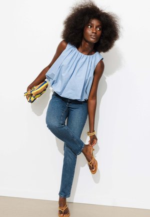 Femme aux cheveux afro ajustant la bride de sa sandale, portant un haut sans manches bleu clair, un jean bleu, tenant une pochette tissée rayée.