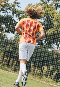 Maglia da calcio arancione con motivo ondulato rosso, pantaloni corti bianchi e scarpini neri. Giocatore con capelli ricci che corre su un campo erboso vicino alla porta.