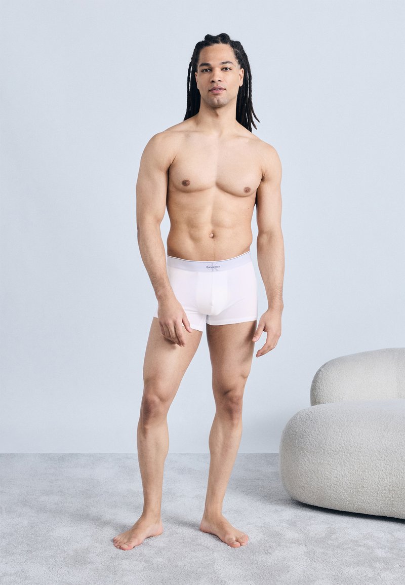 Μυώδης άνδρας με μακριά πλεγμένα μαλλιά, όρθιος ξυπόλητος με λευκά ανδρικά εσώρουχα τύπου μποξέρ, μπροστά σε ανοιχτό γκρι φόντο, με μια καρέκλα με υφή κοντά του.