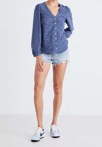 Femme portant un chemisier bleu à motifs floraux boutonné, un short en denim clair déchiré et des baskets blanches, debout devant un fond uni.