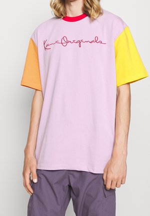 Person iført en løs lyserød T-shirt med orange og gule ærmer, rød krave og rød "Kani Originals" skrift på brystet, kombineret med lilla bukser.