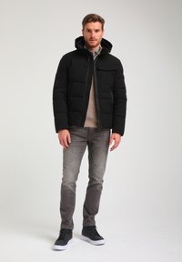 Sort pufferjakke med hætte, lynlås og frontlomme. Bæres over en lys sweater og grå jeans. Sorte sneakers fuldender outfittet.