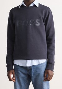 Marinblå sweatshirt med glänsande "BOSS"-logotyp, ribbade muddarna, rund halsringning, lager på lager över en ljusblå skjorta med rutmönster, tillsammans med blå jeans.