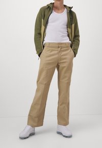 Hombre con una sudadera con cremallera verde y beige, camiseta sin mangas blanca, pantalones beige y zapatillas blancas, de pie con las manos en los bolsillos.