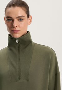 Olivgrön zip-up sweatshirt med hög krage, med ribbad textur och diagonalt sömdesign. Mjuk tyg, minimalistisk look.