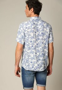 Korte mouwen shirt gemaakt van lichtgewicht stof, met een witte achtergrond en een blauw palmbladpatroon. Ggedragen met denim shorts.
