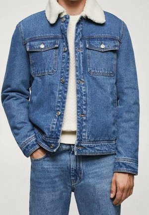 Mann trägt blaue Jeansjacke mit weißem Sherpa-Kragen über weißem Strickpullover und blauen Jeans.