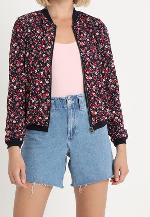 Kvinde iført en sort blomsterprintet bomberjakke med lynlås, lyserød top og højtailede lyseblå denimshorts med frynsete kanter.