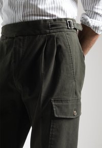 Pantalones caquis hechos de tejido duradero, con un frente plano, pliegues, un bolsillo lateral con botón y un cinturón ajustable elegante.