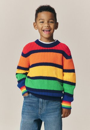 Lachende jonge jongen met handen in de zakken, gekleed in een kleurrijk gestreepte trui en blauwe jeans tegen een effen achtergrond.