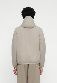 Doudoune beige clair avec capuche, poignets et ourlet élastiques, présentant une texture lisse et un design minimaliste, vue de l'arrière.