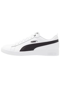 Chaussures de sport blanches en cuir synthétique avec des accents noirs et une bande latérale proéminente. Elles présentent un bout rond, des lacets plats et une semelle texturée.