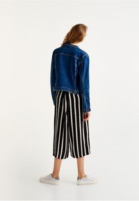 Femme debout, de dos, portant une veste en jean bleue, un pantalon large à rayures noires et blanches, et des baskets blanches.