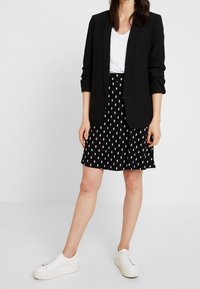Blazer noir à manches longues, associé à une jupe à motifs noir et blanc. Des baskets blanches complètent la tenue. Éléments de design simples et modernes.