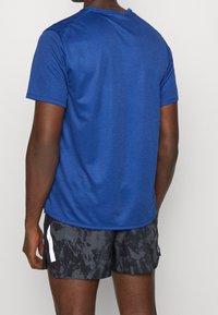 Man som bär en blå kortärmad skjorta och svarta mönstrade träningsshorts med en vit reflekterande rand på sidan, vänd bort.