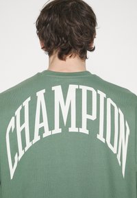 Zelená mikina s nápisem "CHAMPION" velkými bílými písmeny na zádech. Vyrobena z měkké látky s žebrovaným výstřihem.