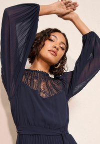 Granatowa sukienka z długimi plisowanymi rękawami, koronkowym detalem w górnej części i dopasowanym paskiem w talii. Teksturowany materiał elegancko kontrastuje z przezroczystymi elementami.