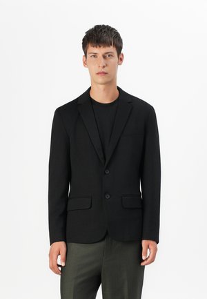 Blazer noir avec revers en pointe, deux boutons et poches avant ; design ajusté associé à un t-shirt sombre et un pantalon olive.
