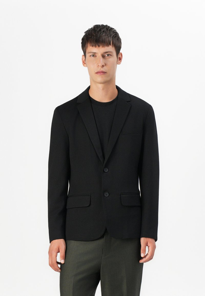 Sort blazeren med almindelige revers, to knapper og forlommer; slim fit design parret med en mørk t-shirt og olivenfarvede bukser.