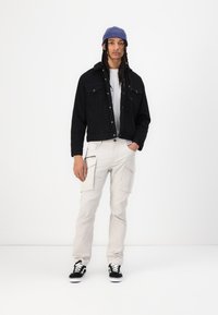 Musta corduroy-takki, jossa on tekoturkiskaulus, valkoinen t-paita, beige cargo-housut sivutaskuilla ja mustat sneakersit valkoisilla pohjilla.