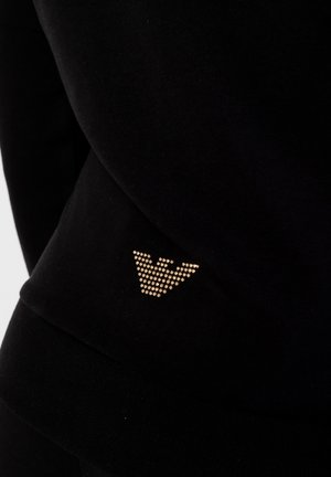 Sweatshirt noir en tissu texturé, avec un accent logo en forme triangulaire clouté d'or près de l'ourlet inférieur.