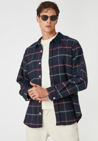Camisa de cuadros en tonos navy con líneas rojas y blancas, cierre frontal con botones y mangas largas, fabricada en una suave tela texturada.