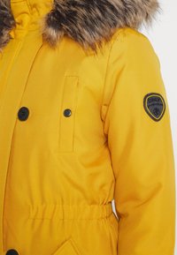 Chaqueta acolchada amarilla con un cuello de piel sintética, cierres de botón negros, cintura elástica y una insignia en la manga. Textura suave.