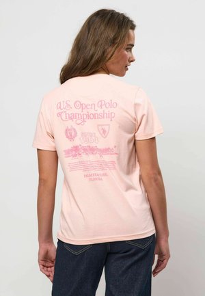 Žena s dlhými vlasmi, ktorá nosí svetloružové tričko s červeným textom a grafikou týkajúcou sa Majstrovstiev USA v póle, v kombinácii s tmavými džínsami.