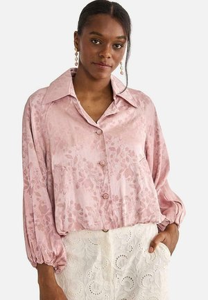 Mujer con una blusa de satén rosa claro con estampado floral y mangas abullonadas, y una falda blanca de encaje, de pie con una mano en el bolsillo.