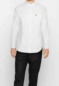 Vit skjorta med lång ärm, i bomull, med en bröstficka och en liten grön logga. Kombination med svarta byxor.