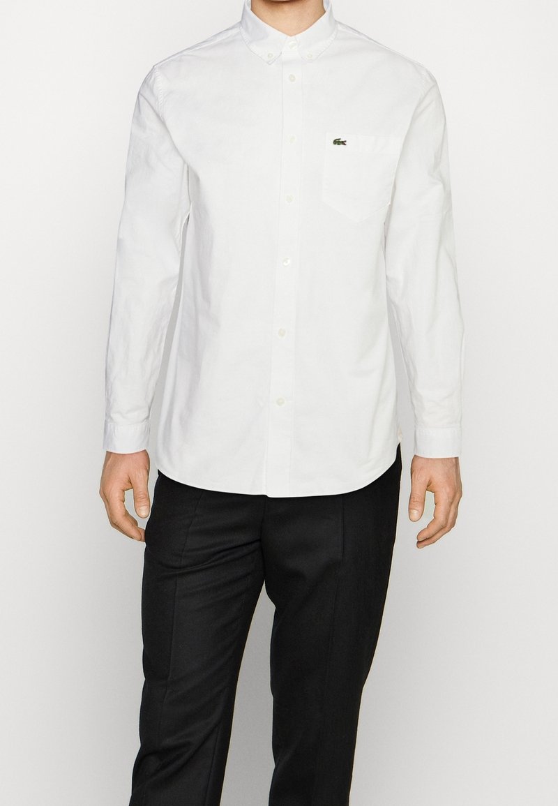 Vit skjorta med lång ärm, i bomull, med en bröstficka och en liten grön logga. Kombination med svarta byxor.