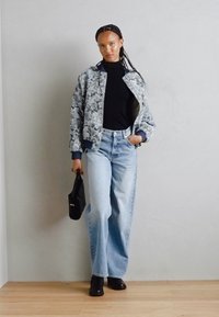 Chaqueta bomber texturizada en gris con un patrón floral, cuello alto negro, jeans de corte alto en azul claro y botines negros.