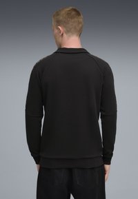 Une personne avec les cheveux blonds courts porte un sweat à capuche noir vu de dos, mettant en avant des embellissements détaillés aux épaules.