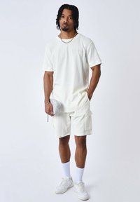 Camiseta blanca texturizada y pantalones cortos a juego, con un bolsillo en los pantalones, combinados con zapatillas blancas y una pequeña bolsa gris.