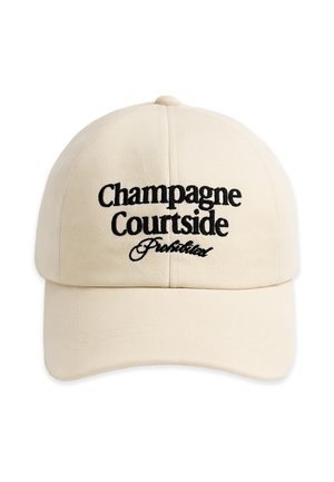 Junger Mann mit hellbeiger Kappe bestickt mit „Champagne Courtside Prohibited“, trägt schwarzes Hemd und graue Jacke über den Schultern, neutraler Hintergrund.