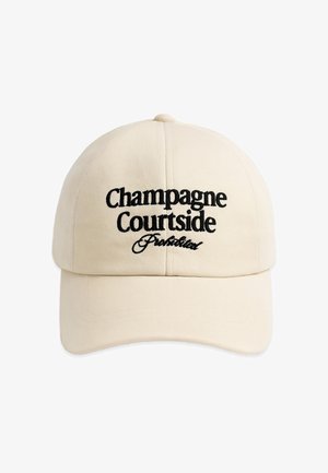 Junger Mann mit hellbeiger Kappe bestickt mit „Champagne Courtside Prohibited“, trägt schwarzes Hemd und graue Jacke über den Schultern, neutraler Hintergrund.