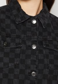 Close-up of black and dark gray checkered denim jacket with silver snap buttons and chest pockets, worn by a person.