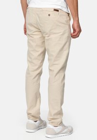 Pantalons beige clair avec une coupe droite, en coton, dotés de poches arrière et d'une étiquette en cuir marron. Les chaussures sont blanches avec des accents gris.