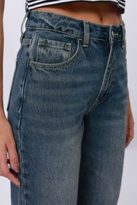 Ženska nosi izbledele modre hlače z visokim pasom in črn-bel navihan top, ki razkriva trebušček ter levo roko z nudastim lakom na nohtih.