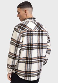 Chemise à carreaux en tons blancs, noirs et oranges. Fabriquée en tissu texturé avec un design à boutons et un ourlet arrondi. Présente un empiècement dans le dos.