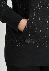 Main dans la poche avant d'un sweat à capuche noir avec de petits motifs fléchés bleus et blancs et des poignets et un bas côtelés.