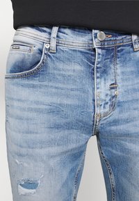 Jeans de denim azul claro con un aspecto descolorido, que cuentan con un cierre de botón frontal, cinco bolsillos y detalles desgastados en el muslo izquierdo.