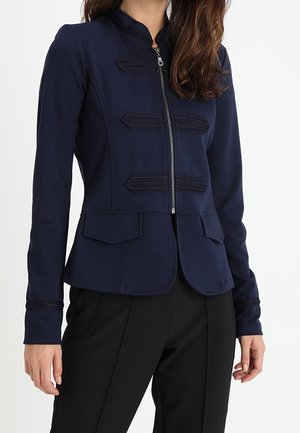 Frau trägt einen taillierten marineblauen Blazer mit Reißverschluss, dekorativem geflochtenem Besatz und Pattentaschen, kombiniert mit schwarzen Hosen.