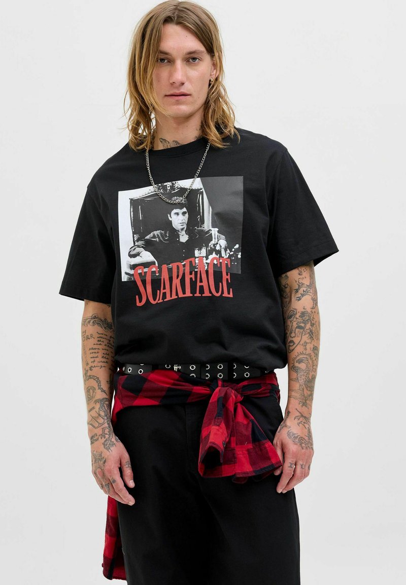 Sort T-shirt med et grafisk print af en karakter og "SCARFACE" i røde bogstaver. Bæreren har en rød- og sortternet skjorte bundet omkring livet.