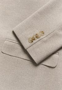 Beige tailored jacket featuring a textured fabric, peaked lapel, and four light brown buttons on the cuff with visible stitching details.