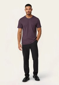 Man staat in een donkerpaars shirt met korte mouwen, zwarte broeken en zwarte schoenen tegen een lichte achtergrond.