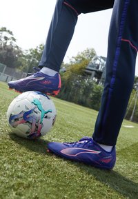 Lilla fotballsko med rosa detaljer og strukturert overflate som hviler på en multicolor fotball, plassert på grønt gress.