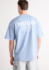 Ljust blå kortärmad t-shirt med vitt logotyp "HUGO" och text på ryggen. Mjukt bomullstyg med avslappnad passform.