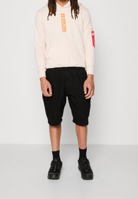 Jasnoróżowa bluza z kapturem z napisem "ALPHA", czarne szorty cargo z bocznymi kieszeniami oraz czarne buty sportowe noszone z czarnymi skarpetami.