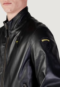 Giacca in pelle nera con colletto alto, chiusura con zip anteriore e toppe sulle spalle con il logo "Blauer". Texture liscia con un design aderente.